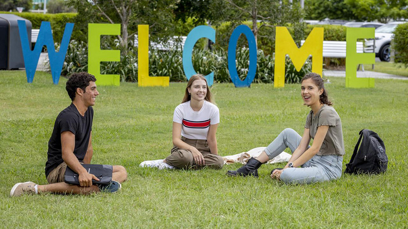 three students sitting in front of a large colourful welcome sign