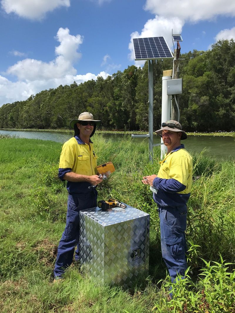 Two men in fluro yellow work clothes next to water monitoring equipment