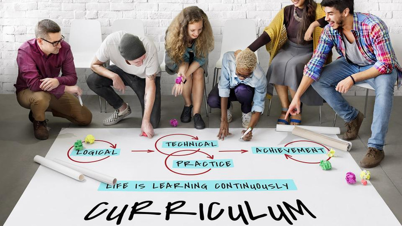 six people sitting along a bench surrounding a large poster of paper on the ground where they are drawing elements of curriculum design